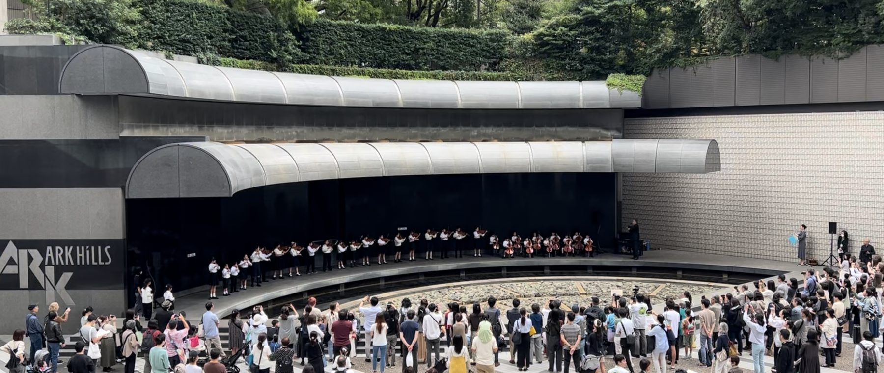 半円の屋根の下で子どもたちが並んで演奏し、広場で大勢が聴いている