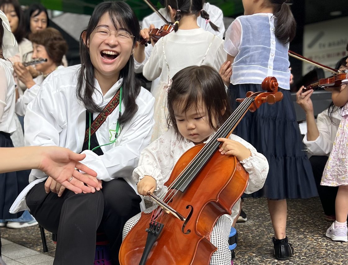 笑顔の先生のそばで、小さな子がはじめてチェロを構えている